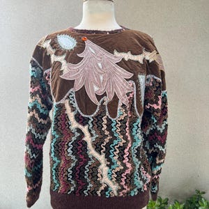 Può includere: Un maglione marrone con un motivo colorato e testurizzato e un grande disegno ricamato sul davanti. Il disegno presenta un albero rosa con accenti dorati e bianchi.