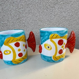 May include: Two ceramic mugs shaped like fish with blue, yellow, red, and white paint. The mugs have a white interior and are designed to look like a tropical fish.