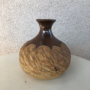 May include: A small, brown and tan ceramic vase with a textured surface. The vase has a rounded body and a narrow neck.