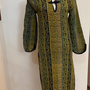 May include: A long-sleeved, floor-length dress with a V-neck and black trim. The dress features a repeating pattern of green, yellow, and black vertical stripes. The dress is displayed on a white mannequin.