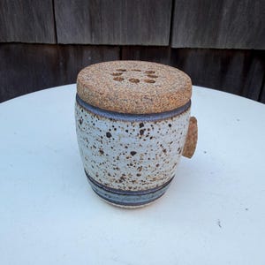 May include: A ceramic salt or spice cellar with a speckled white and brown exterior. The cylindrical container has a cork lid with three small holes and a small cork handle. A blue band encircles the base and top of the container.