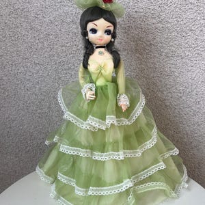 May include: A vintage doll with dark hair and large blue eyes, wearing a green dress with multiple ruffled layers and white lace trim. The doll has a green hat with a red rose and a black choker.