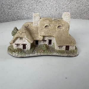 May include: A small, detailed model of a stone cottage with a thatched roof, two chimneys, and a small tree in front. The model is made of a light brown material and is sitting on a white surface.