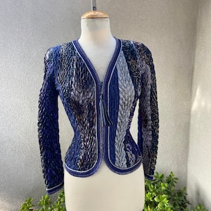 Puede incluir: Una chaqueta azul y plateada con cuentas y cierre de cremallera. La chaqueta tiene una silueta ajustada y una tela texturizada.