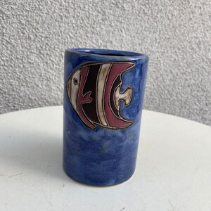May include: A blue ceramic mug with a white and pink fish design. The mug is glazed and has a rustic look.