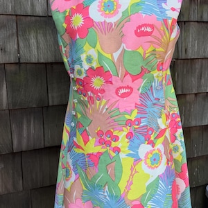 Puede incluir: Un vestido vintage con un estampado floral vibrante en rosa, amarillo, verde y azul. El vestido sin mangas presenta un escote redondo y una fila de botones perlados en el lateral. El vestido se exhibe en un maniquí.