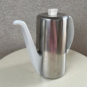 May include: A silver teapot with a white spout and handle. The teapot has a white lid with a small knob on top.