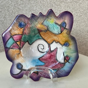May include: A small, colorful ceramic dish with an abstract design. The dish is painted in shades of purple, pink, blue, green, and yellow. It has a white interior and a slightly raised rim.