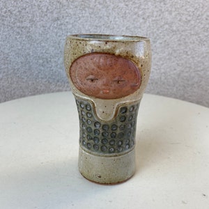 May include: A small, ceramic vase with a brown face and a green and white speckled pattern. The vase is handcrafted and has a unique, whimsical design.