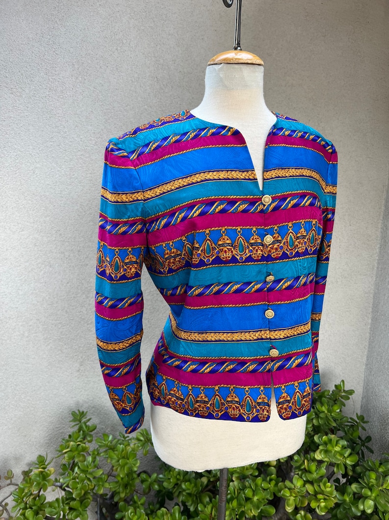 May include: A long-sleeved, button-up blouse with a colorful striped pattern. The pattern features blue, turquoise, pink, gold, and purple stripes with a repeating design of gold chains and jeweled pendants.