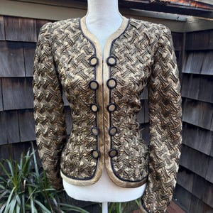 May include: Gold and black textured jacket with a round neckline and long sleeves. The jacket features black buttons and trim, and is displayed on a white mannequin. The jacket's design is intricate and detailed, with a woven pattern.