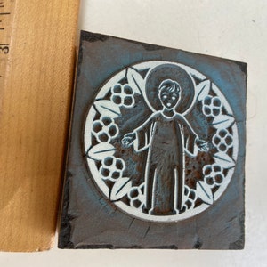 May include: A vintage woodblock printing stamp with a blue and white design of a child in a halo with a floral border.