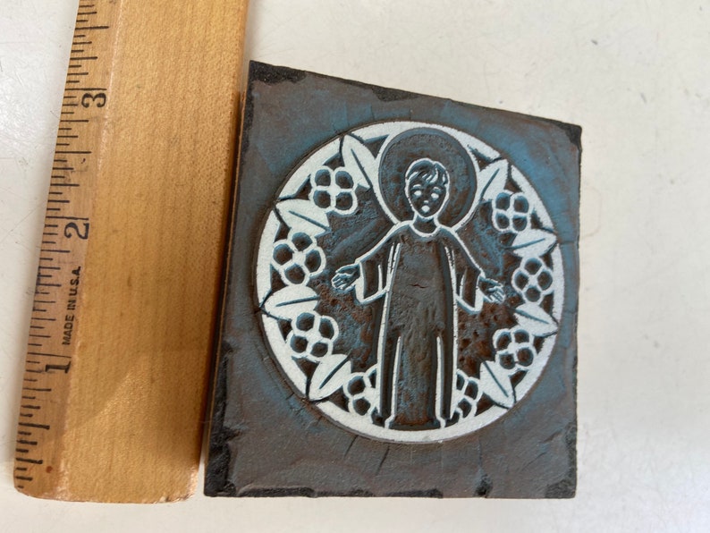 May include: A vintage woodblock printing stamp with a blue and white design of a child in a halo with a floral border.