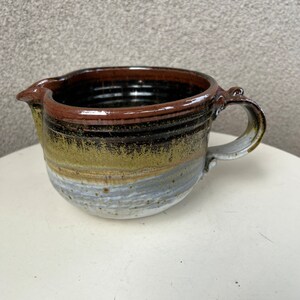 May include: A brown, black, and green ceramic pitcher with a white glaze. The pitcher has a wide, round base and a handle on the side.