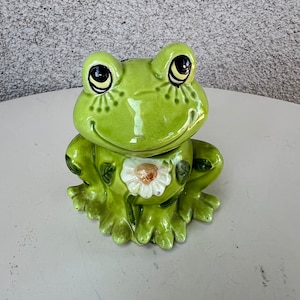 Può includere: Una statuetta di rana in ceramica verde con un fiore bianco e foglie verdi sul petto. La rana ha grandi occhi neri e un ampio sorriso.