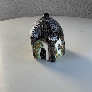 May include: A small ceramic cottage with a thatched roof, white walls, and a green and brown ivy design. The cottage has a small door and a window.