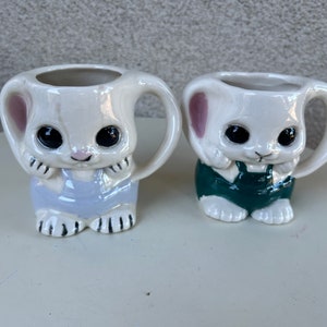 May include: Two white ceramic bunny mugs with black eyes and pink ears. One mug has a light blue and white outfit and the other has a green and white outfit.