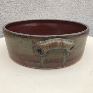 May include: A handmade ceramic bowl with a dark brown interior and a muted green exterior. The bowl features a unique handle design on each side. The handles are a lighter brown with blue accents. The bowl is a round shape.