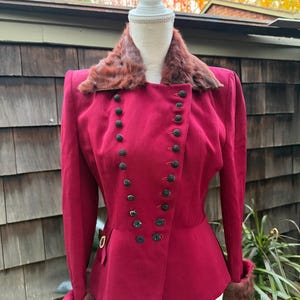 Può includere: Giacca bordeaux con colletto e polsini in pelliccia, esposta su un manichino. La giacca presenta un design doppiopetto con bottoni neri e una vita aderente. Lo sfondo comprende un muro di legno e fogliame.