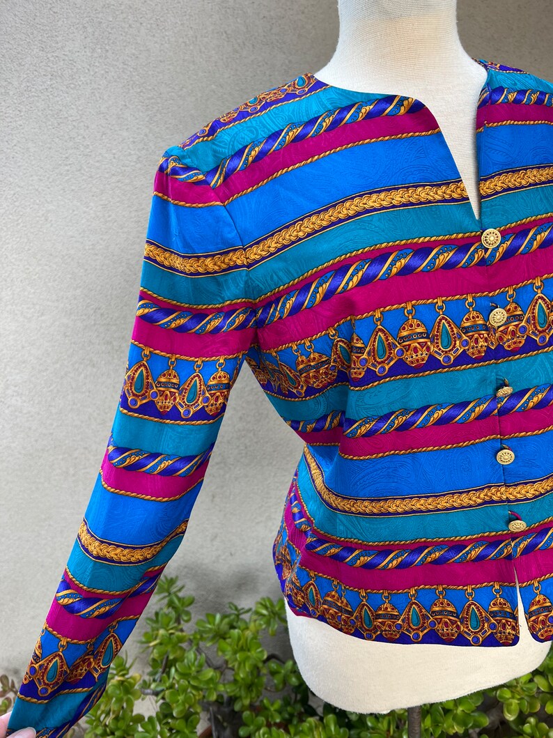 May include: A long-sleeved blouse with a colorful striped pattern featuring gold chains, turquoise, pink, and blue. The blouse has a V-neck and button closure.