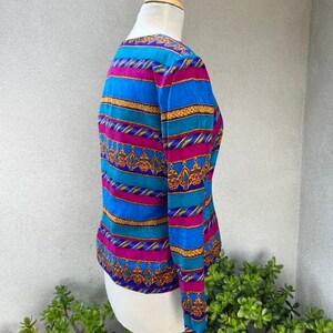 May include: A long-sleeved blouse with a colorful striped pattern. The stripes are blue, teal, pink, and gold. The pattern includes a repeating design of gold chains and dangling jewels.