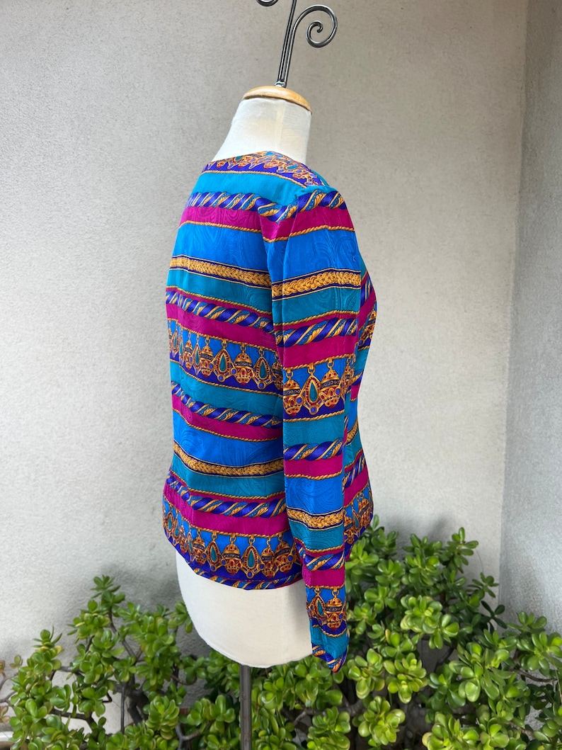 May include: A long-sleeved blouse with a colorful striped pattern. The stripes are blue, teal, pink, and gold. The pattern includes a repeating design of gold chains and dangling jewels.
