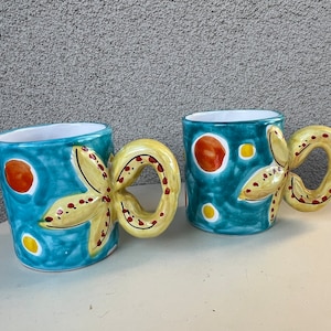 May include: Two blue ceramic mugs with starfish handles. The mugs are decorated with yellow starfish, red dots, and yellow dots on a blue background.