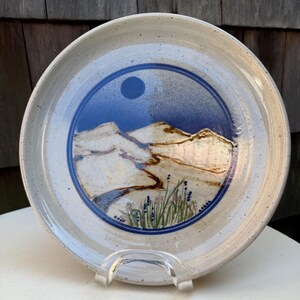 May include: A round ceramic plate with a speckled off-white glaze. The center features a circular blue inlay depicting a landscape with rolling hills, a dark blue sun, and green and purple wildflowers in the foreground, all under a clear glaze.