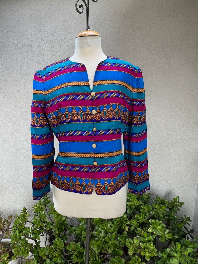 May include: A long-sleeved, button-up blouse with a colorful striped pattern. The fabric features a geometric design with gold accents. The blouse has a V-neck and a fitted silhouette.