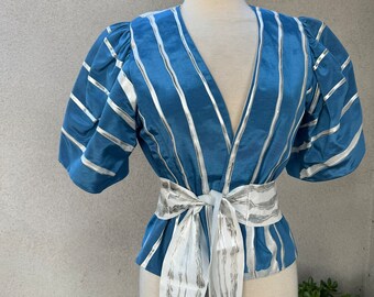 May include: A blue and silver striped blouse with a white sash tied at the waist. The blouse has a V-neckline and short, puffed sleeves.