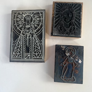 May include: Three vintage wood block printing stamps. One features a religious figure with a halo and a child, another depicts a face with a radiant halo, and the third shows a religious figure with a halo.