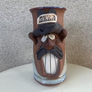 Puede incluir: Una jarra de cerveza de cerámica marrón con un diseño de cara sonriente. La jarra presenta un bigote negro, un sombrero negro con la palabra "BEER" y una boca abierta blanca. La jarra tiene un borde azul y marrón.