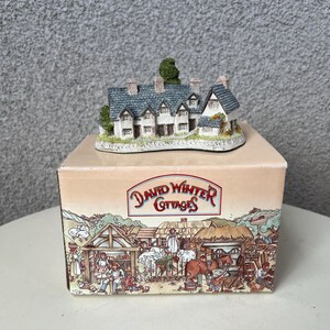 May include: A white cottage with a grey roof and green trees, part of a David Winter Cottages collection. The cottage is on a base with a scene of a village with people and animals.