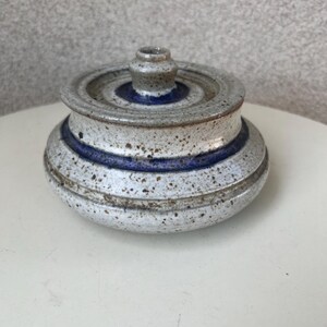May include: A speckled ceramic jar with a lid. The jar is primarily white with a blue band and a small knob on the lid. The jar has a rustic, handmade appearance, suitable for kitchen or home decor.