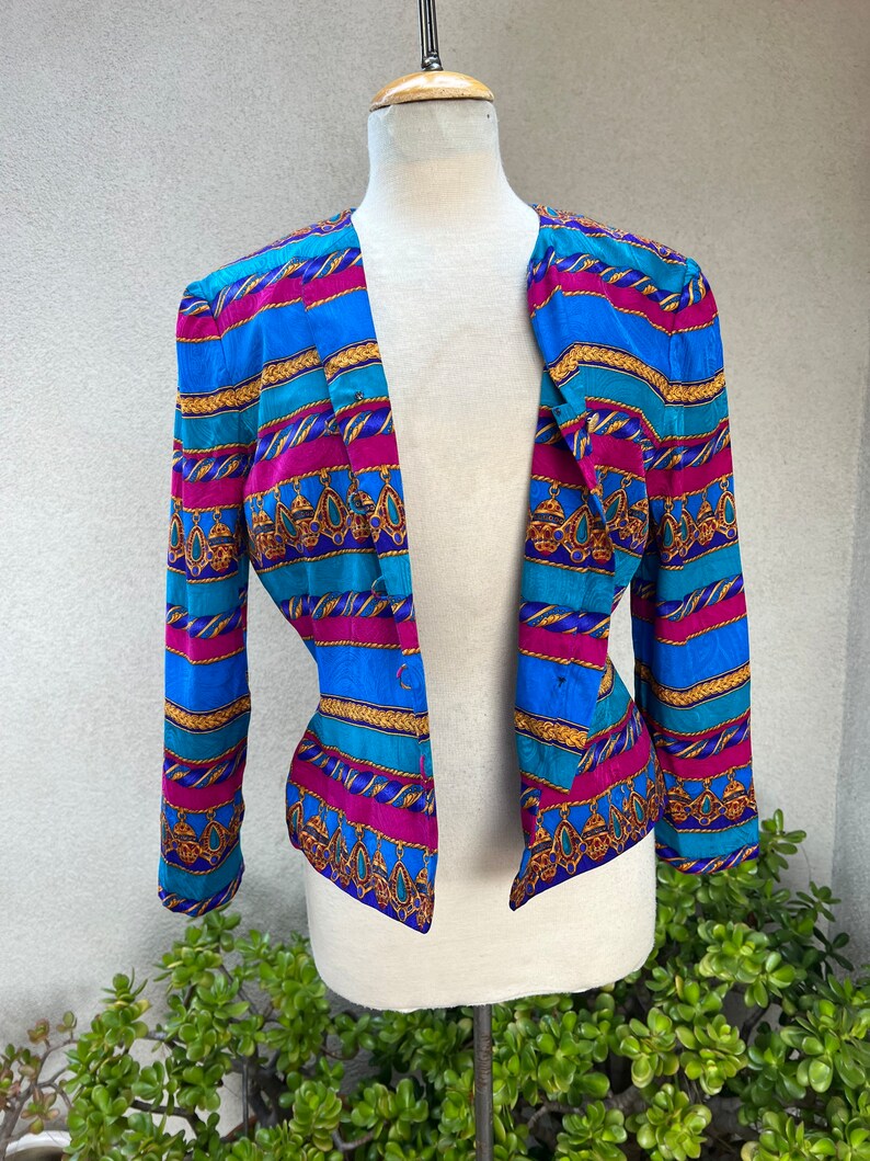 May include: A blue, pink, and gold striped blazer with a geometric pattern. The blazer has a long sleeve and a button closure.