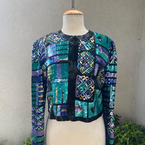 Può includere: Giacca corta con paillettes dal design patchwork. La giacca presenta un mix di paillettes turchesi, viola e blu con finiture nere. Le maniche sono lunghe e la giacca è esposta su un manichino.