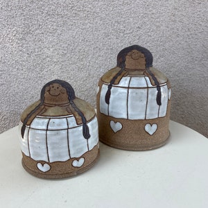 Puede incluir: Dos jarras de cerámica con un diseño pintado a mano de una niña sonriente con cabello castaño y un vestido blanco. Las jarras tienen una base marrón con tres corazones blancos.