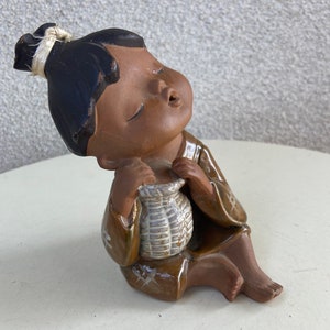 Może przedstawiać: Brązowa ceramiczna figurka osoby z czarnymi włosami, ubrana w brązową szatę i trzymająca koszyk. Figurka siedzi ze skrzyżowanymi nogami i odchyloną głową do tyłu.