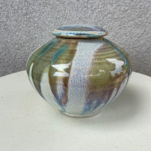 Könnte beinhalten: Eine Keramikdose mit Deckel, die eine weiße Basis mit grünen, blauen und braunen Streifen aufweist. Die Dose hat eine abgerundete Form und eine glatte, glänzende Oberfläche.