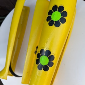 May include: Bright yellow wooden leg supports with black flower designs and green centers. The supports are curved and appear to be part of a larger piece of furniture. The background is a white surface.