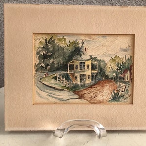 May include: A watercolor painting of a yellow house with a white picket fence and a winding road leading to it. The house has a pointed roof and a porch. The painting is framed in a cream-colored mat.