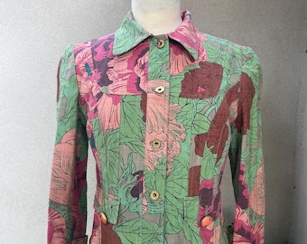 May include: A floral print jacket in shades of green, pink, and brown, displayed on a mannequin. The jacket features a collared neckline, long sleeves, and gold-colored buttons. The fabric has a patchwork design with large floral patterns.