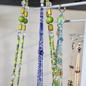 May include: A collection of beaded necklaces and earrings in various colors. The necklaces feature green, yellow, and clear beads, while others are blue or turquoise. Some earrings have paw print charms.