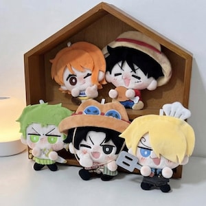May include: Five plush dolls representing anime characters are arranged on wooden shelves. The dolls feature distinct hair colours including orange, black, green, and yellow, and wear various accessories like hats and a spatula, all with embroidered faces.
