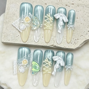 May include: A set of ten artificial nails with a light blue and cream color scheme. Each nail features decorative elements such as lace, butterflies, bows, and corset-style lacing. The nails are arranged on a neutral surface.