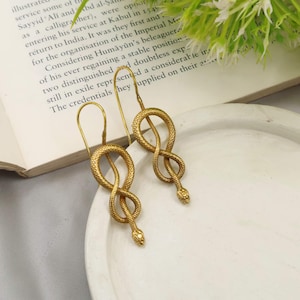 May include: A pair of gold-toned drop earrings featuring a textured snake design. Each earring is shaped into a looped figure-eight pattern with a detailed serpent head at the bottom. The earrings hang from long, curved wire hooks for secure placement.
