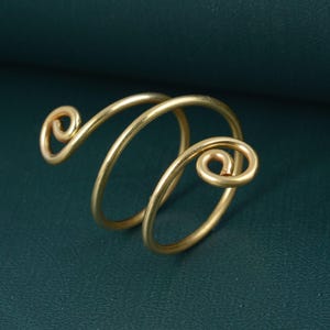 Könnte beinhalten: Ein goldfarbener Ring mit einem Spiraldesign. Der Ring hat ein durchgehendes, gebogenes Band, das sich um den Finger wickelt, mit dekorativen Wirbeln an jedem Ende. Der Ring besteht aus einem glänzenden, metallischen Material.