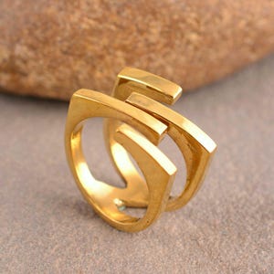 May include: A close-up shot of a gold-colored ring with a unique, geometric design. The ring features a series of connected rectangular and curved elements, creating an abstract shape. The ring is made of a shiny, metallic material, reflecting light.