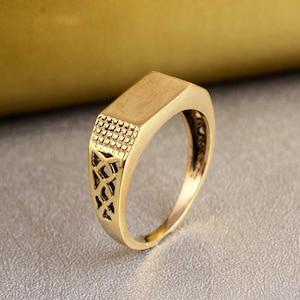 May include: A gold-toned ring featuring a flat, rectangular top surface. The band displays a textured grid pattern near the head, transitioning into an openwork, lattice-style design along the sides of the shank, creating a detailed metal structure.