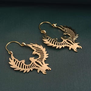 May include: A pair of gold-coloured hoop earrings. Each earring features a stylised phoenix design with intricate cut-out details. The phoenix is depicted with outstretched wings and a detailed tail. The earrings are displayed against a dark green background.